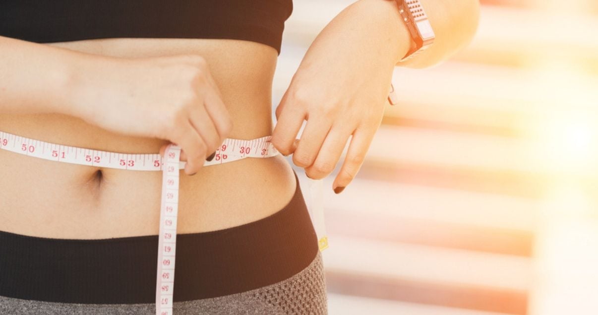 Mujer joven adulta vestida con indumentaria deportiva y un reloj para tu optimizar su rendimiento durante su entrenamiento físico, utiliza una cinta metrica para medir su cintura y asi complementar su calculo de imc